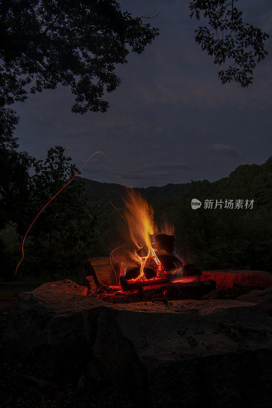 长时间暴露的夜晚篝火的火焰和弯弯曲曲的余烬路径在左，与烟雾山天际线在背景