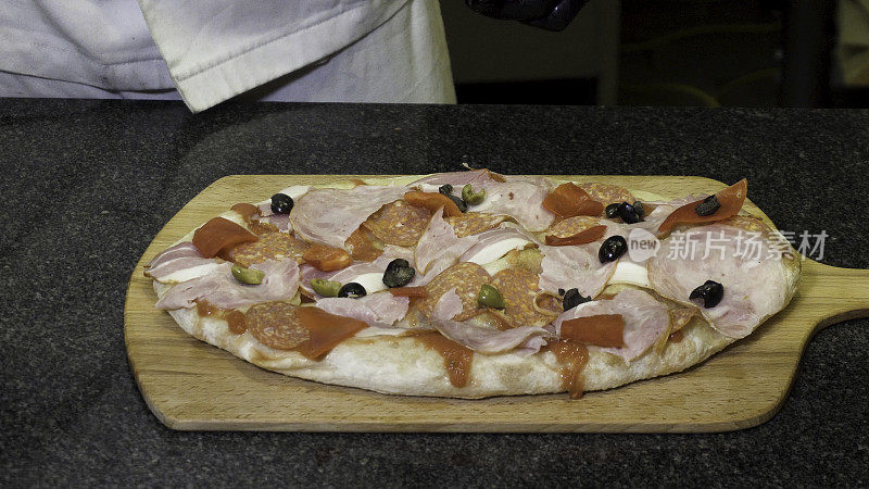 穿着黑色烹饪手套的厨师躺在木板上制作披萨，美味的食物概念。框架。手把橄榄和肉放在披萨上的特写。