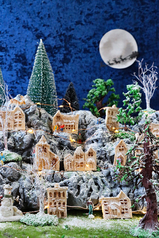 风景优美的圣诞村展示的图像，人造塑料和木制圣诞树在山上设置，照明自制的木屋，圣诞老人雪橇和驯鹿飞过满月的剪影，重点在前景