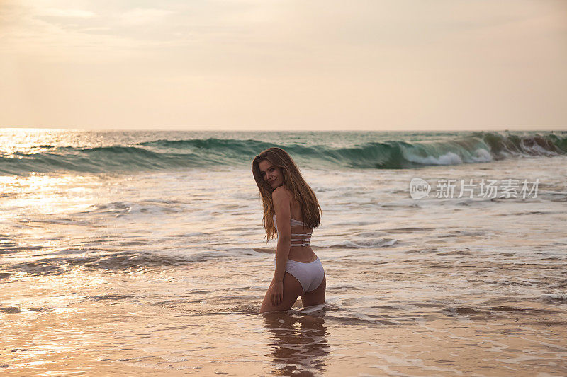 年轻苗条的女人在白色泳装摆姿势在海水背景，看着相机的背影。轻松愉快的热带假期。旅游度假度假概念。复制广告文字空间