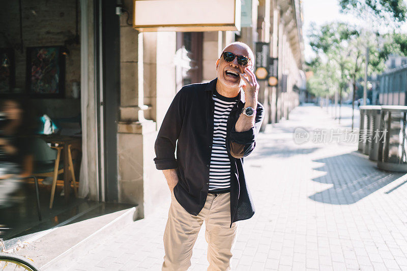 有趣的男性游客戴着太阳镜，在现代智能手机设备上使用漫游互联网连接讨论退休后的国际度假，快乐的老人在城市里一边打电话一边笑