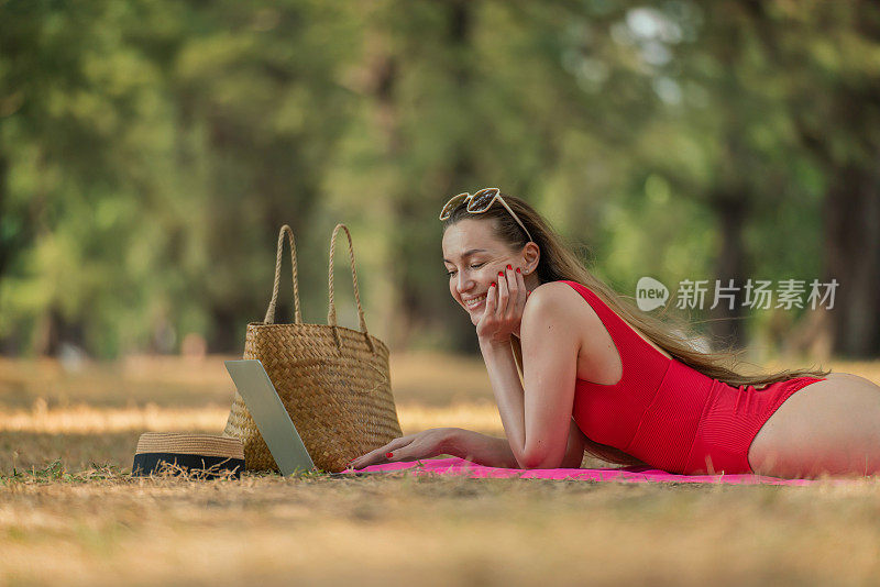 美丽的白人女性性感的女人在红色泳装比基尼戴着太阳镜躺在地上工作在她的电脑笔记本电脑放松在树林里的海滩