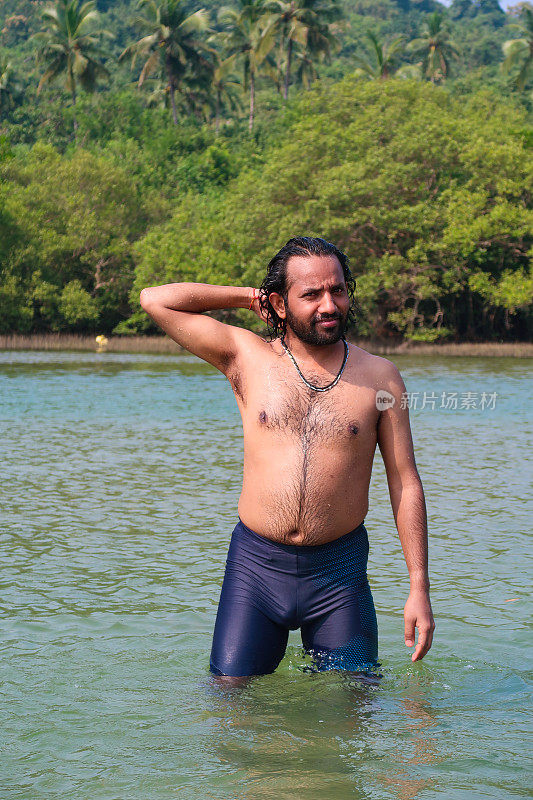 特写图片:穿着冲浪短裤的印度男子从平静的泻湖中涉水而出，用手拂去脸上湿漉漉的头发，热带岛屿天堂，有棕榈树的林地景观，重点在前景