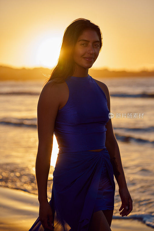 美丽的拉丁美洲女人在日落时穿着蓝色连衣裙微笑着走在海滩上