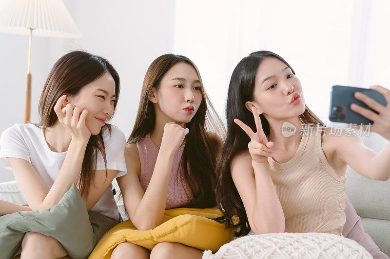 三位年轻的亚洲千禧一代女性坐在家里客厅的沙发上聊天，用智能手机自拍。最好的朋友，友谊，相聚的时光，科技的理念。