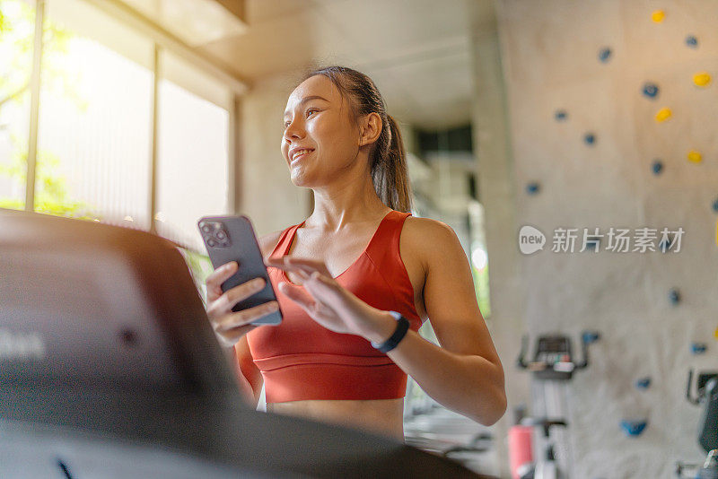 一名亚洲女性在室内健身俱乐部的阳光下用智能手机在跑步机上休息