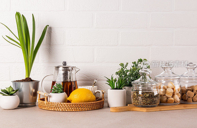 时尚的厨房背景与玻璃茶壶，柠檬，杯子，茶罐，糖，蛋白酥皮。花盆里的绿色植物。前视图。白砖墙。