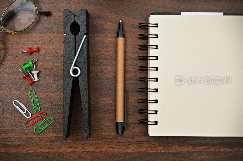 将文具，如钢笔、日记本、螺旋记事本、回形针、别针、眼镜等的水平照片美观地放在深棕色的木质水平背景上。