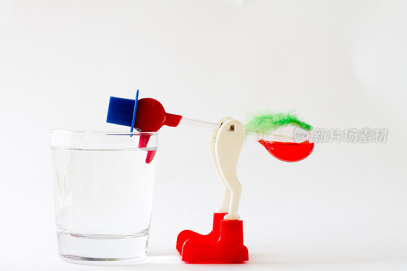 白色背景上的玩具“饮水鸟”。饮水鸟斜靠在一杯水里。