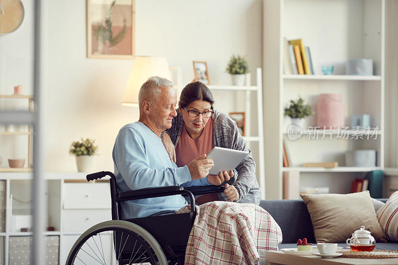 一位腿上盖着毯子的老人坐在轮椅上，向惊讶的妻子展示平板电脑上的新应用程序