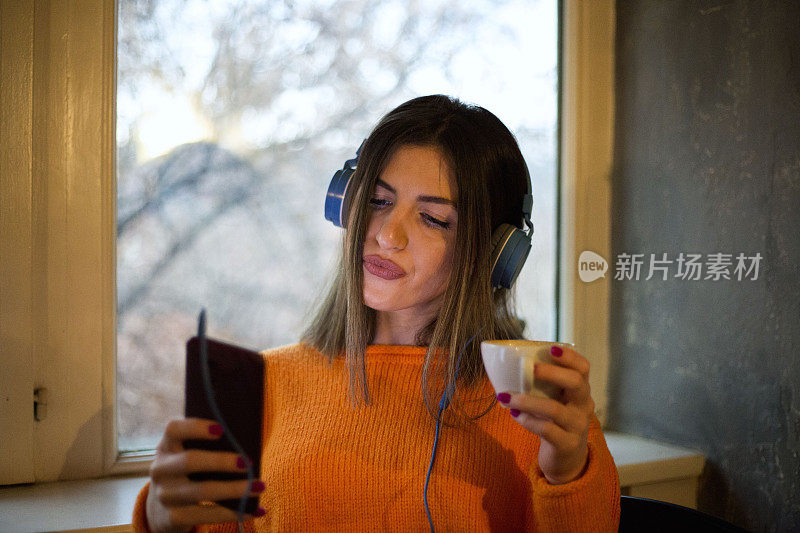 年轻漂亮的女人在窗边喝咖啡，戴着耳机听音乐