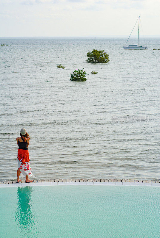 一个女人站在泻湖的无边泳池边上，背景是一艘游艇