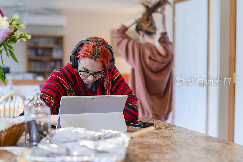 在家学习并不总是舒适的。这个十几岁的女孩戴着耳机在远处上课，在厨房柜台边用笔记本电脑工作，这时她的大姐正从后面走进客厅。