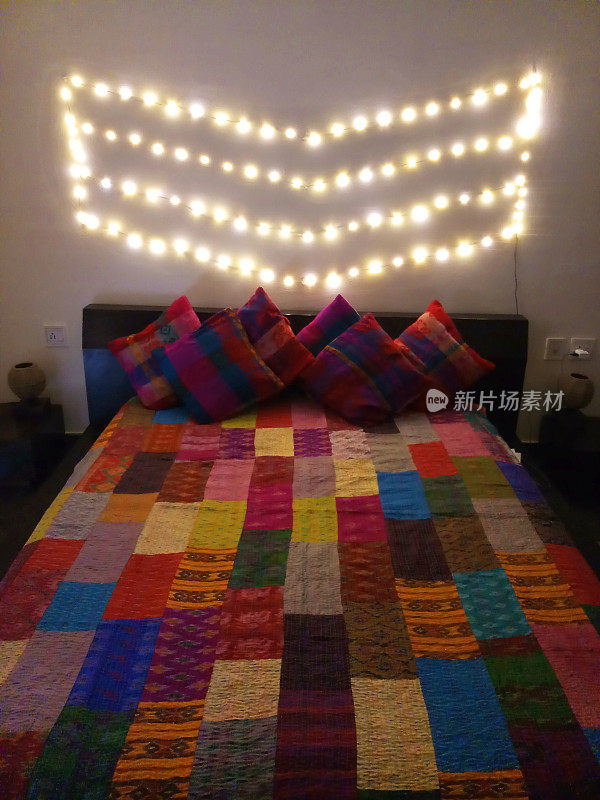 床头板上斜倚着色彩鲜艳的散垫，墙上有灯光，拼花图案设计的被子，五彩缤纷的床罩