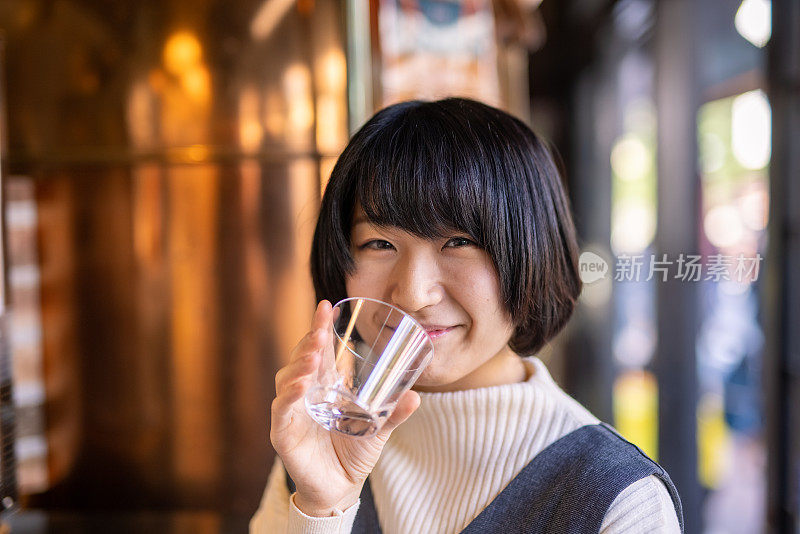 快乐的年轻女子喝一杯“saki”(日本米酒)在酒类商店