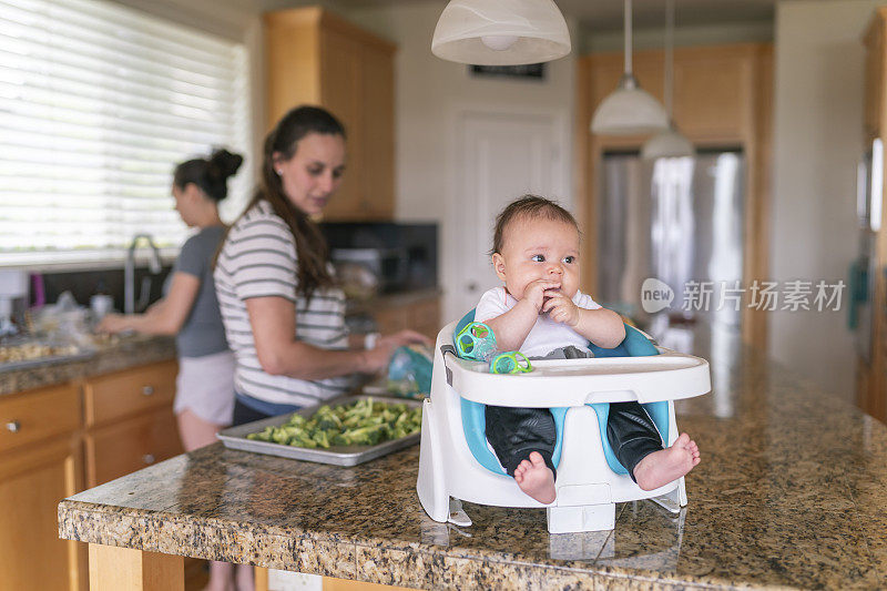 小女孩坐在厨房柜台上的婴儿椅上，其他人在做午饭