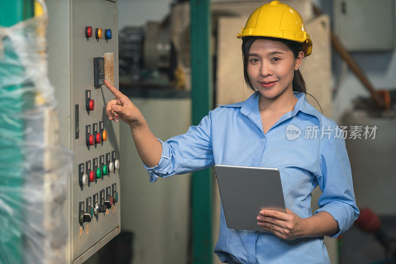 亚洲女性工业工厂工人，戴着黄色安全帽，手拿平板电脑，站在工厂的电子机器控制面板旁工作。工业工厂工人日常工作的概念