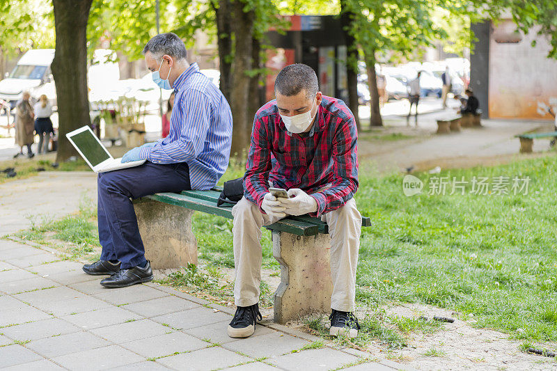 公园里的两名商人使用笔记本电脑进行远程工作，佩戴防护口罩，保持社交距离以避免冠状病毒传播。