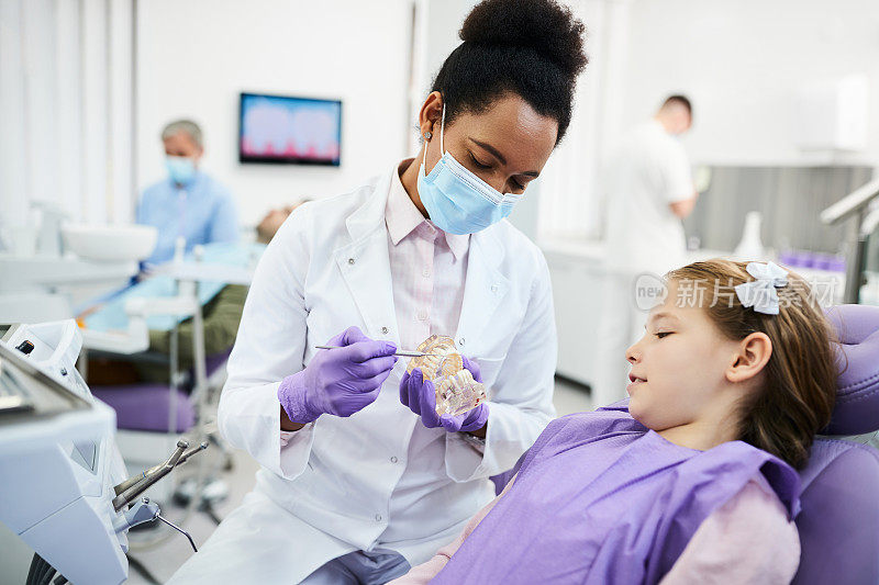 非裔美国牙医使用牙科模型，同时与小女孩在牙科诊所预约。