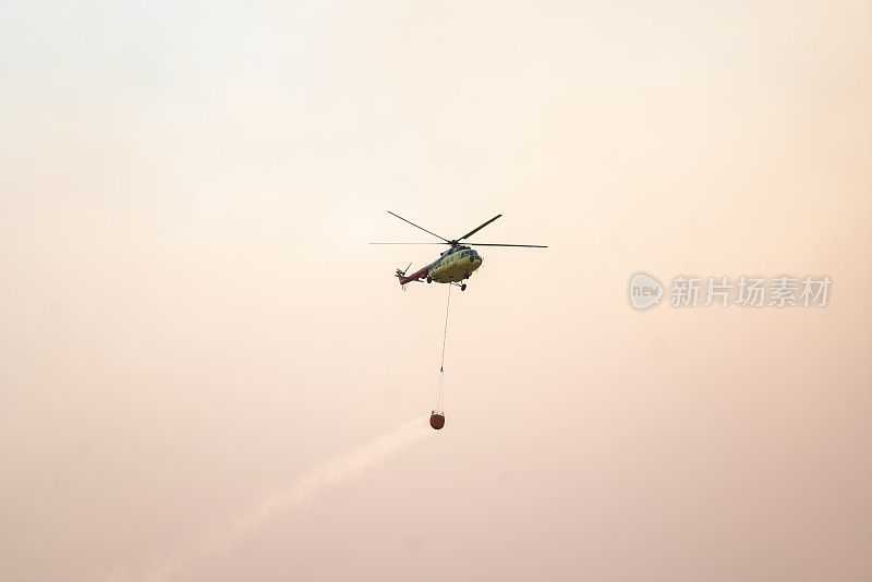 空中消防在黄色多云的天空背景中带着水篮的飞行直升机。