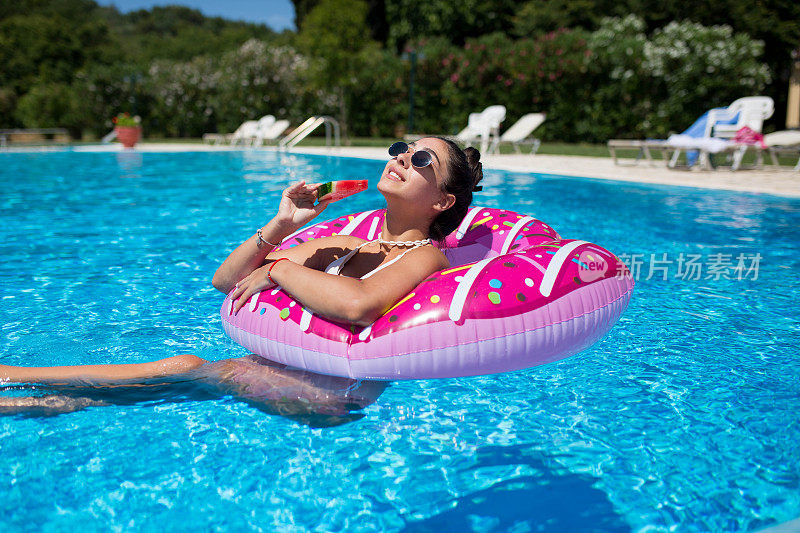 一个美丽的微笑的女孩在游泳池吃西瓜，享受一个炎热的夏天