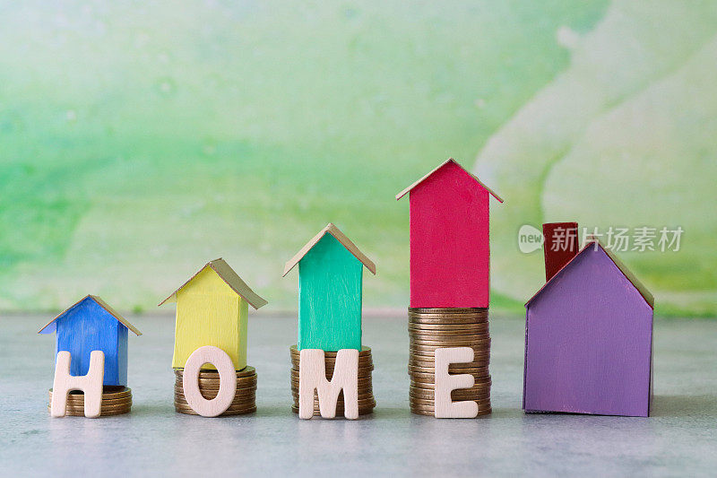 图片是一排排彩色纸板房子，上面是一堆堆硬币，紫色的模型房子旁边是拼写“家”的木制字母，重点是前景，斑纹绿色背景，房地产和储蓄概念