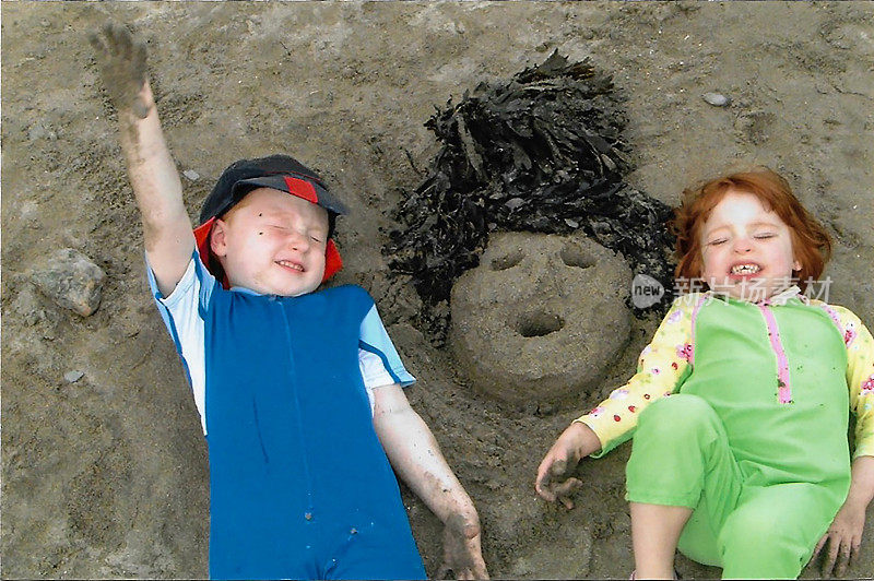 特写图像两个红头发的孩子躺在沙滩上闭着眼睛，穿着防紫外线泳衣，沙脸海藻头发，高高地观看