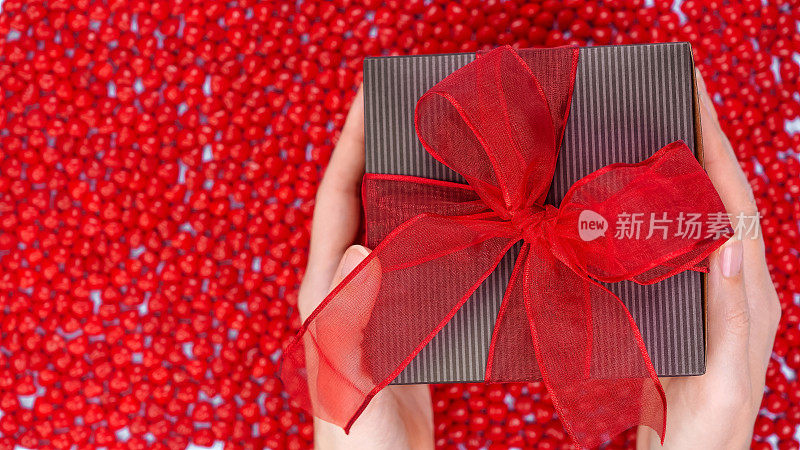 女手持圣诞礼盒装饰红丝带蝴蝶结在红色糖果背景。圣诞和新年横幅。圣诞购物节礼日薄荷糖