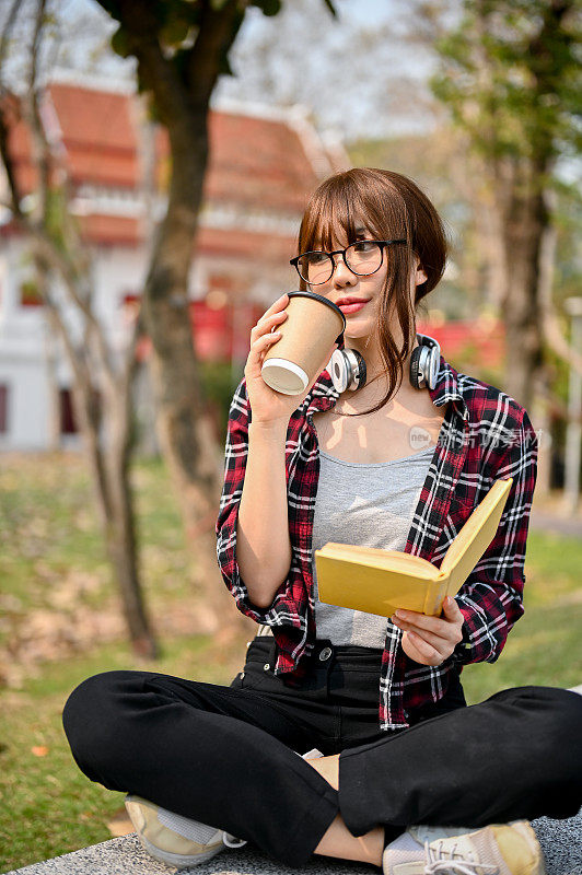 年轻漂亮的亚洲女大学生正在喝咖啡和阅读一本书在长凳上