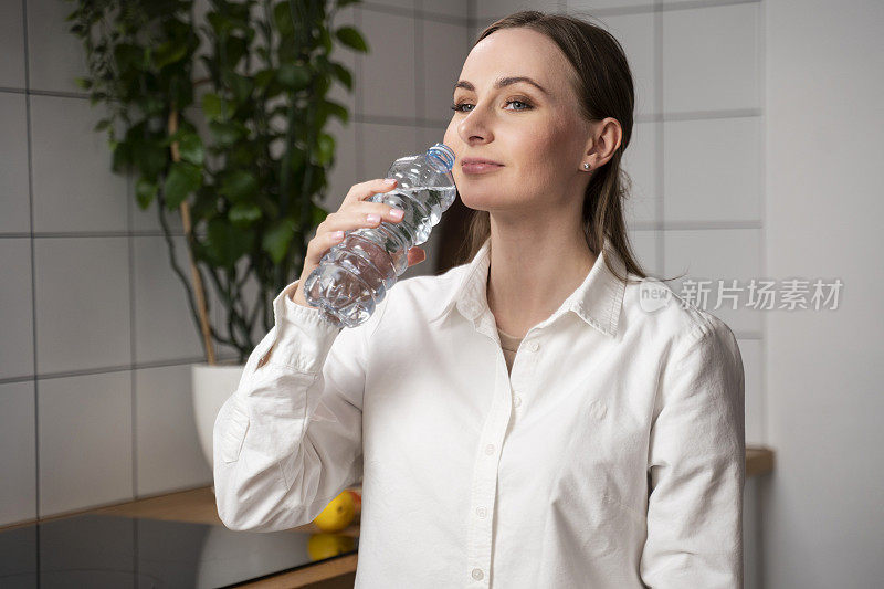 漂亮的年轻女子穿着白衬衫，留着长发，从塑料瓶里喝水，微笑着看着镜头，向镜头展示了一瓶水。