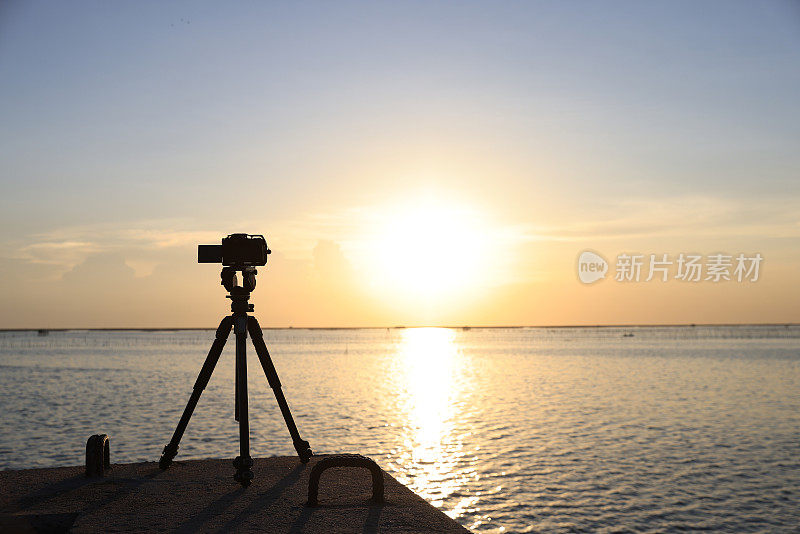 三脚架上的照相机正在拍摄海边的日落。设置一个等待日落的相机，在海边拍一张风景照。