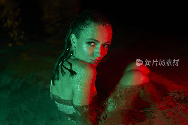 女子晚上坐在水环境中，被绿色和红色的灯光照亮，创造了一个戏剧性的时尚场景。潮湿的女性在水中，生动的色彩反射皮肤在黑暗的氛围创意照片。
