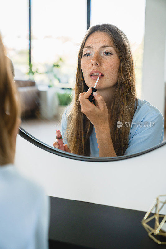 年轻的白人女性在家里对着镜子涂口红的垂直肖像。