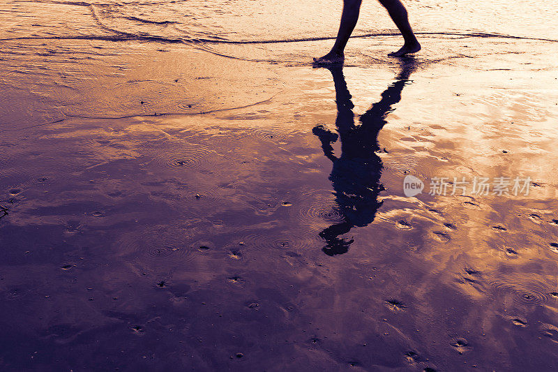 日落时在海边的沙滩上散步的人。倒影在水镜中的剪影。
