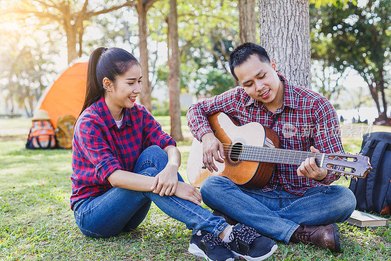 年轻的夫妇露营享受弹吉他和坐在户外露营帐篷，夫妇露营的概念