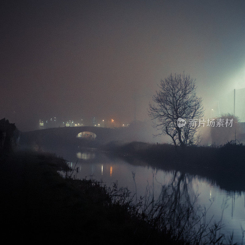 夜晚雾气蒙蒙的运河，树和桥被照亮，阴森恐怖的气氛和平静的运河水