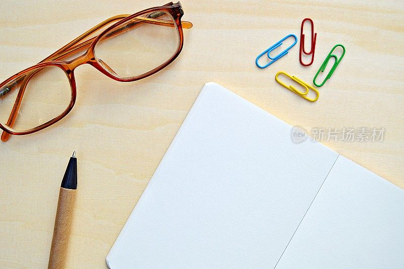 一张水平的照片，一张白色的素色记事本，一个棕色和黑色的钢笔和彩色回形针，眼镜放在一个木制的米色水平背景上，很有美感。