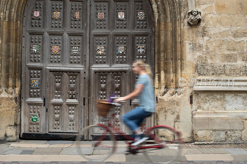 女学生在运动模糊的牛津大学学院大楼周围骑老式自行车