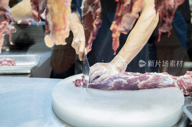 在一家牛肉火锅餐厅的开放式厨房里，厨师在挂着的切碎的生肉后面用刀切牛肉