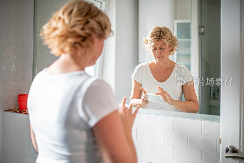 金发女人在浴室镜子前从罐子里取面霜