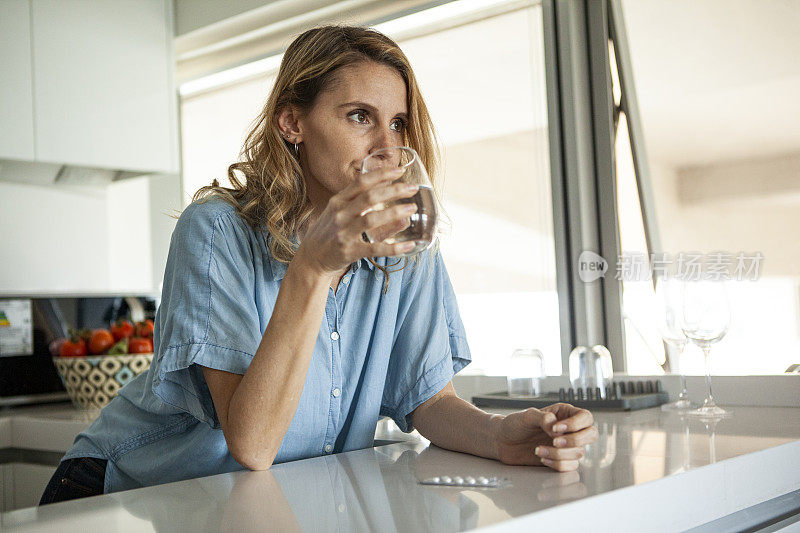 中年妇女拿着一杯水在厨房里吃药丸的肖像