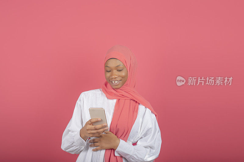 高兴的年轻穆斯林女子用智能手机进行社交，在网上聊天中做出回应，在社交媒体上发布一些东西，穿着粉色面纱和白色衬衫，孤立在粉色背景下。