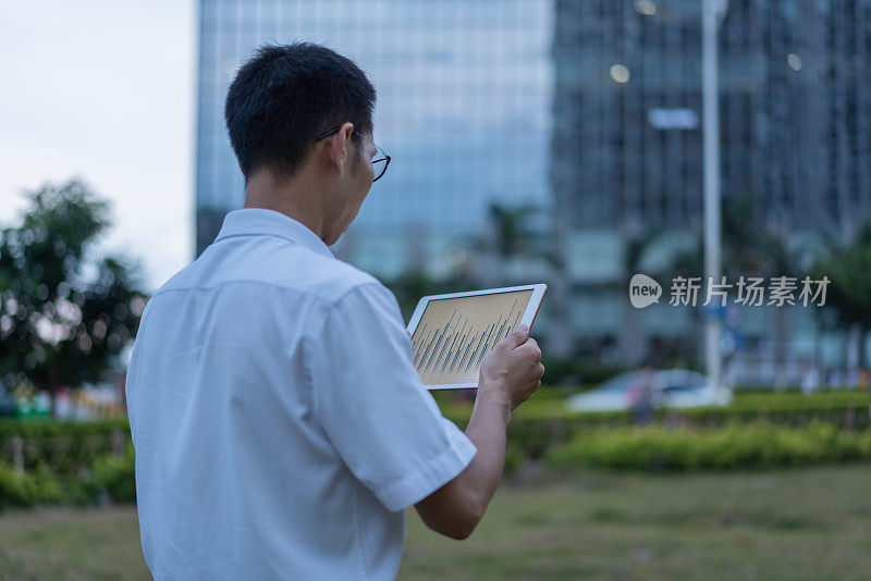 一位亚洲男性白领正在办公大楼外的草地上使用平板电脑