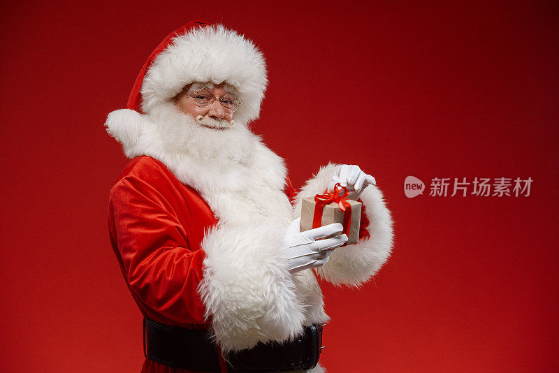 圣诞老人手里拿着一个小礼盒，站在红色的背景上，看着相机