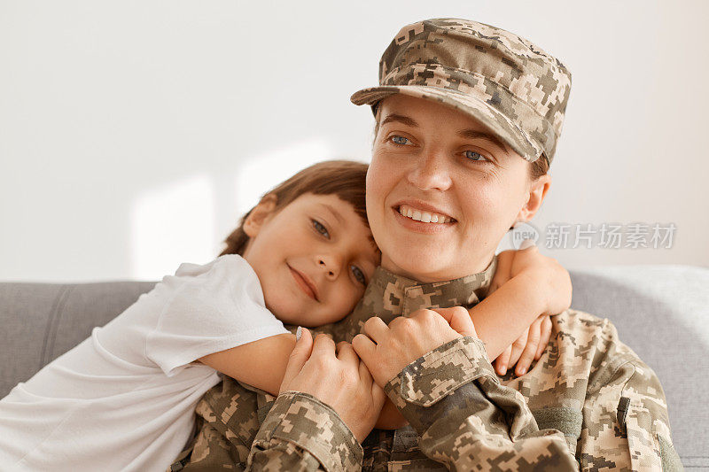 室内拍摄身穿迷彩服、头戴迷彩帽的女军人从战场或军队归来，坐在沙发上抱着孩子，可爱的女儿想念母亲，拥抱母亲。
