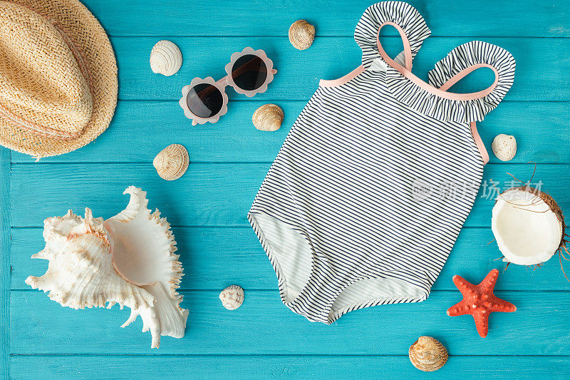 儿童连体泳衣和草帽旁边的海滩配件，贝壳，海星和椰子上的木制蓝绿色背景。夏日氛围和海边度假的概念。平躺