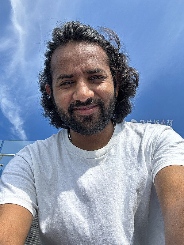 印度男子用手机自拍的特写图像，拿着手机，低头看手机屏幕自拍，阳光明媚的蓝色多云的天空背景，专注于前景