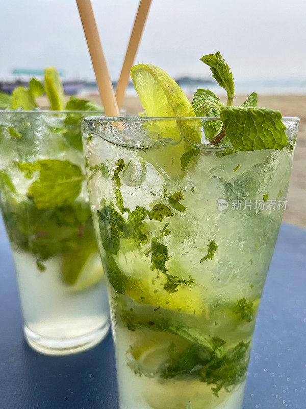 特写图像的海滩餐厅酒吧场景，玻璃杯莫吉托鸡尾酒碎冰，薄荷叶枝，柠檬片和吸管在桌子上，重点在前景