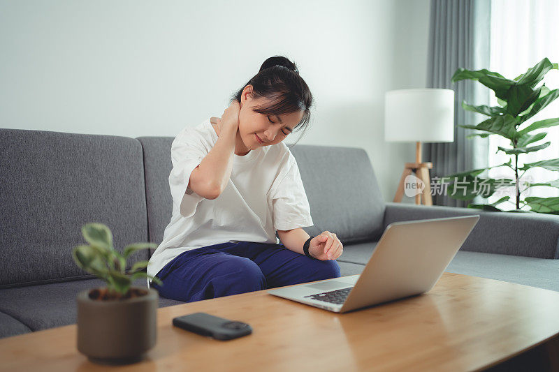 一名亚洲女性坐在家里的沙发上使用笔记本电脑，身体疼痛。