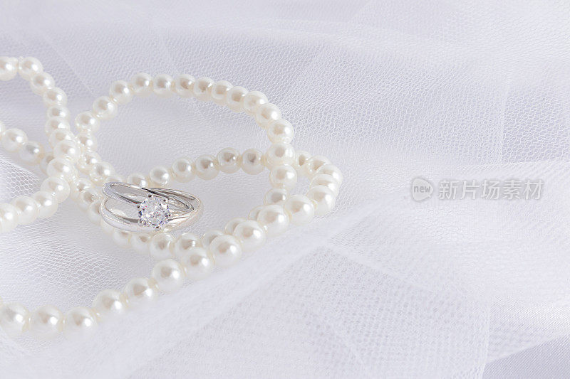 美丽的婚礼背景设计。两枚镶有钻石的白金订婚戒指，戴在精致的白色新娘头纱上，头纱上缀有珍珠珠。